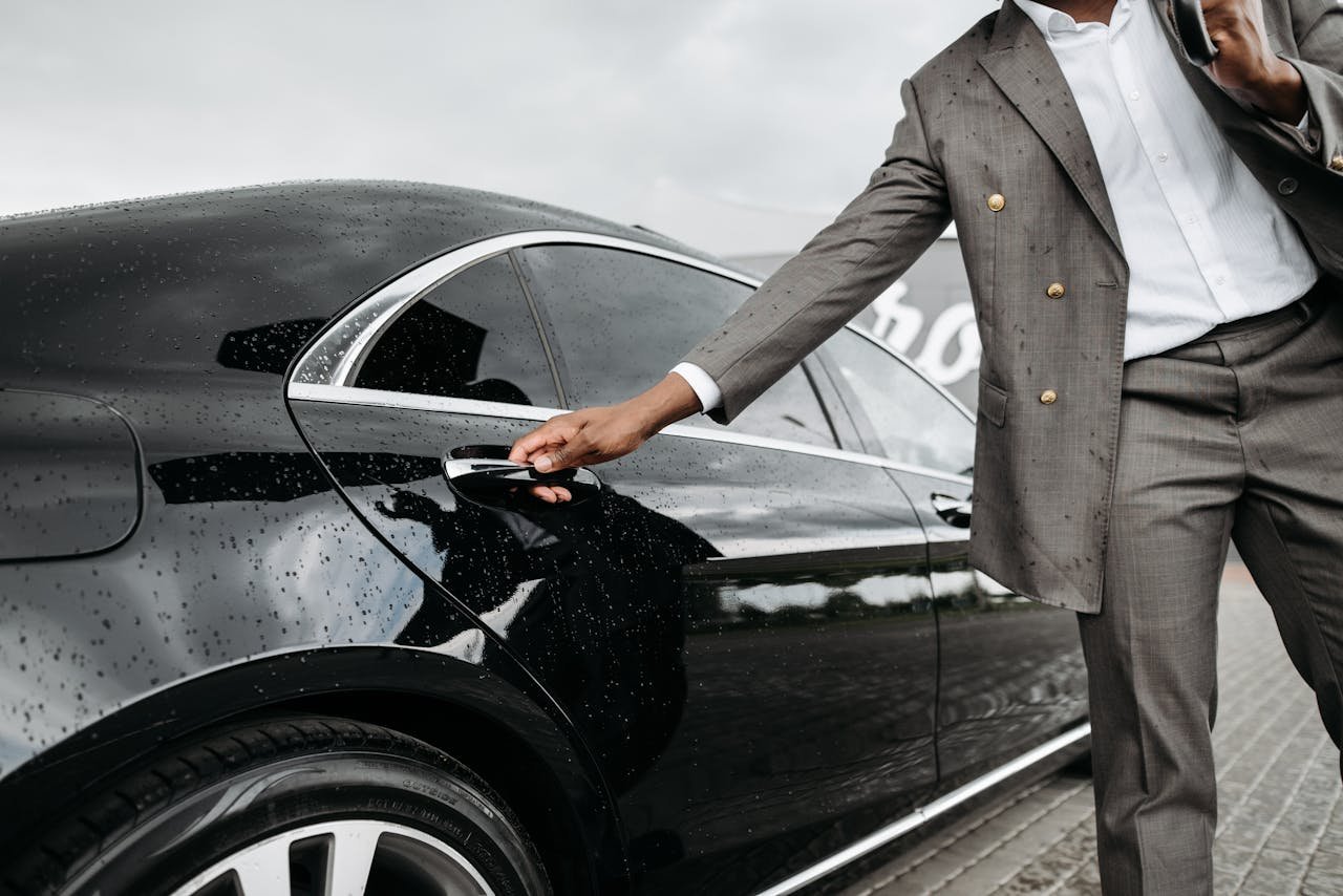 A man in a suit opens a luxury car door under raindrops on stone pavement.